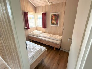 A small, single-room accommodation with a wooden floor, two beds, a window with curtains, and a painting on the wall. - Holiday Home in Otterndorf (Otterndorf)