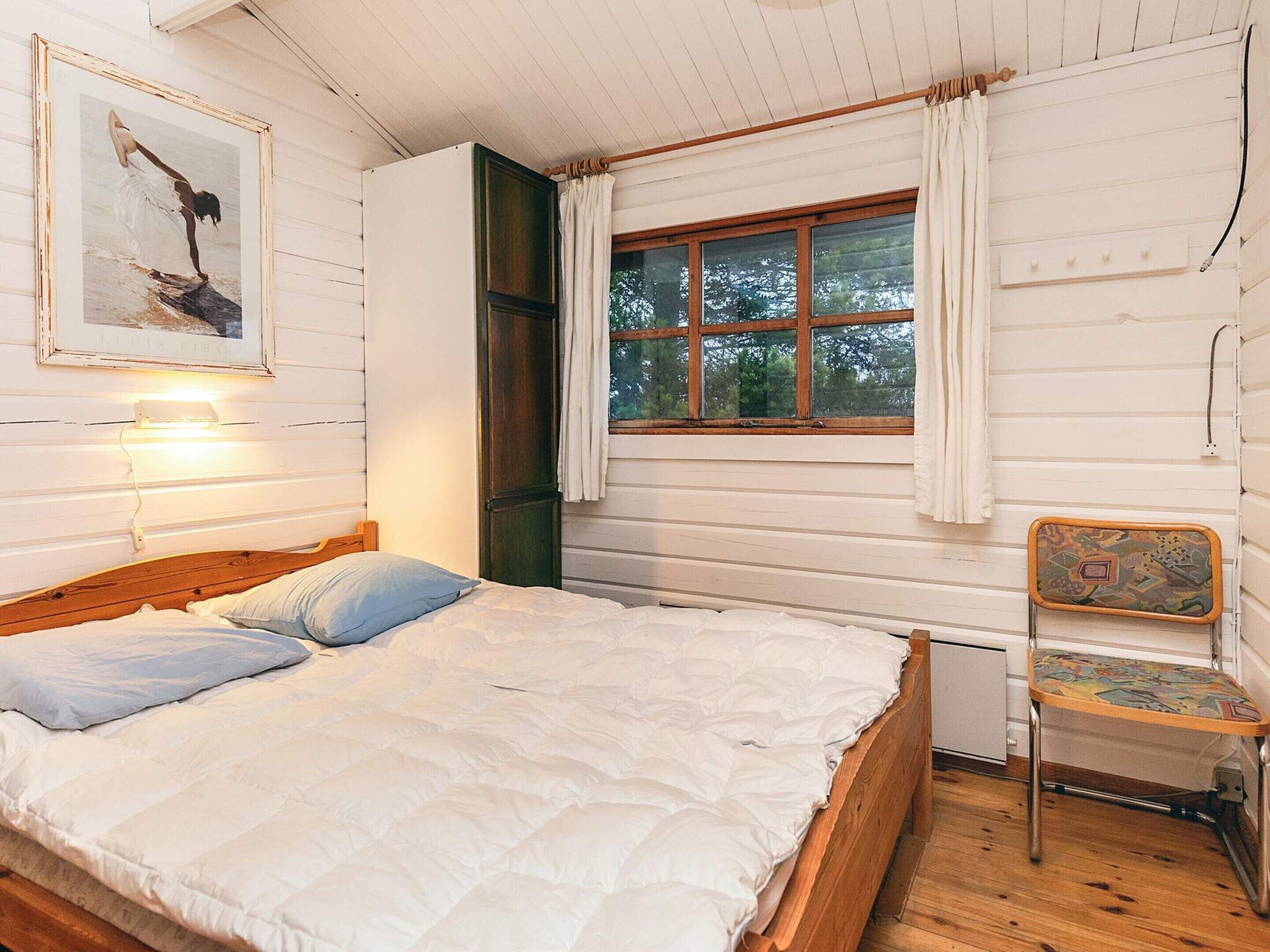 A bedroom with a wooden bed, a window with curtains, a chair, and a wall-mounted light.