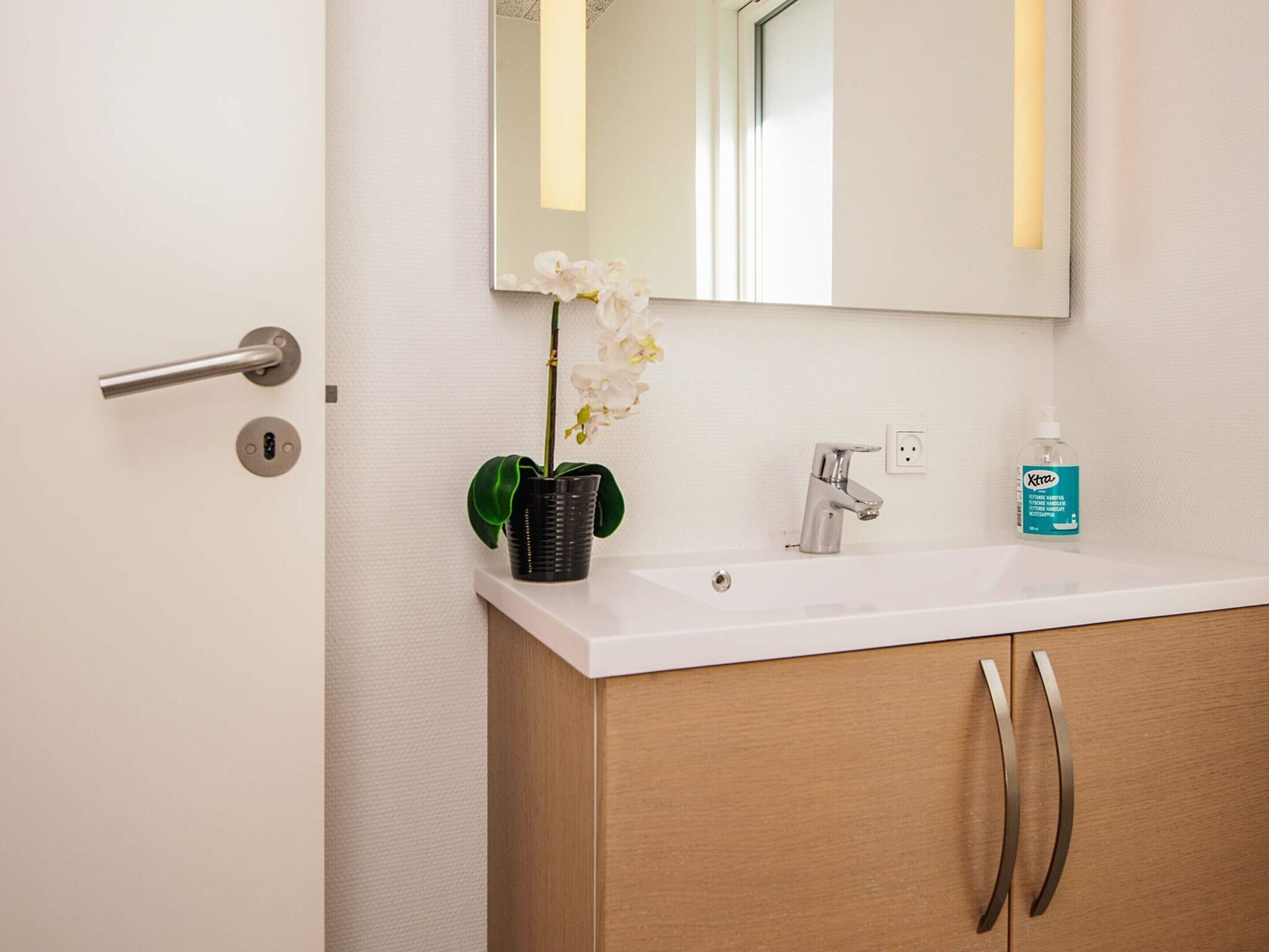 Une salle de bain avec un lavabo blanc, un miroir, une orchidée en pot et un flacon de savon pour les mains.