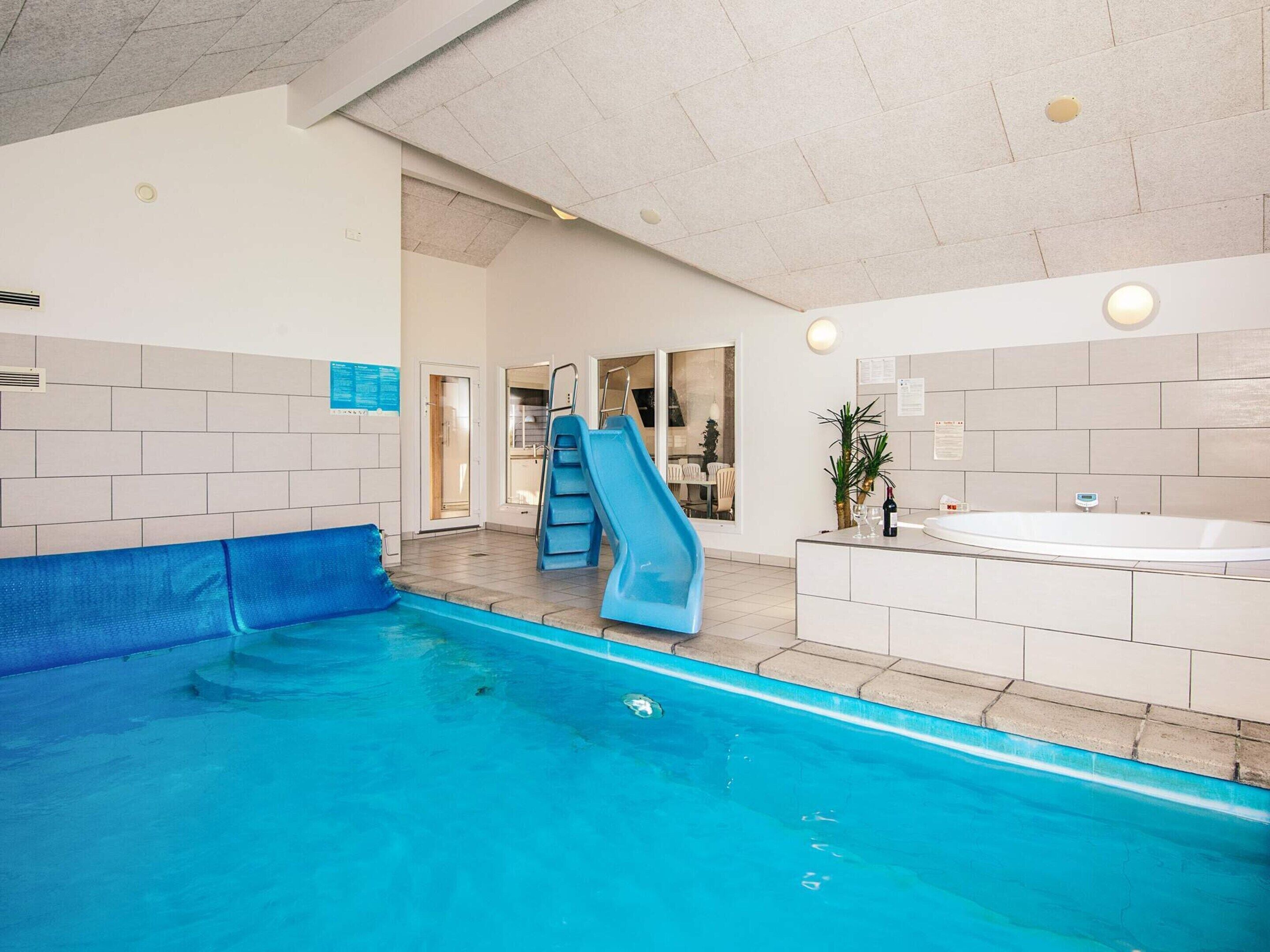 Une piscine intérieure avec un toboggan bleu, un jacuzzi et un escalier menant à un espace baigné de lumière.