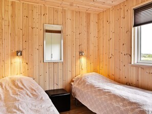 A room with wooden panel walls, two single beds with white linens, a small black bench, a mirror, and a window with blinds.