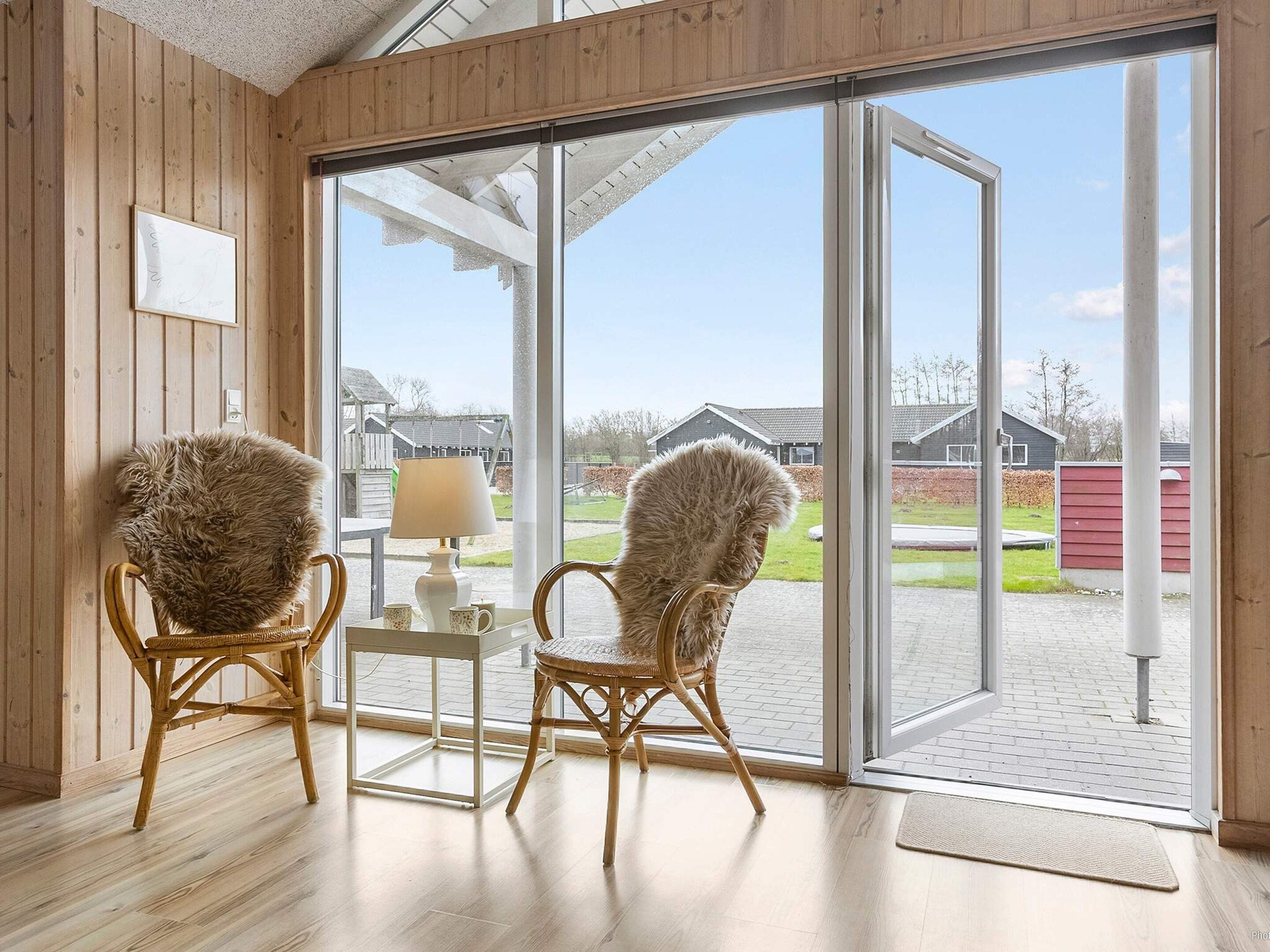 A room with wooden walls, two wicker chairs, a small table with a lamp, and a large glass door leading outside.