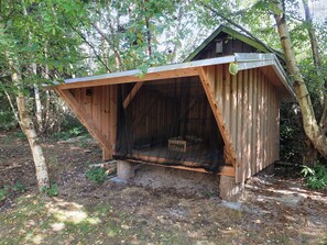 Una estructura de madera con techo inclinado, posiblemente un cobertizo o una cabaña pequeña, rodeada de árboles y vegetación natural.