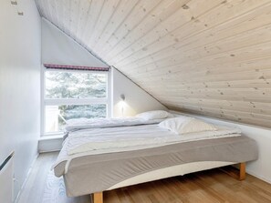 A bedroom with a sloped ceiling, a bed with white bedding, a window with a view of trees, and a wall-mounted light fixture.