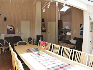 A dining area with a long table, chairs, and a floral centerpiece. There is a kitchen in the background with appliances and a microwave. - Holiday Home in Otterndorf (Otterndorf)