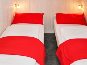 Two single beds with red bedding and white sheets, positioned against a wooden wall with two wall-mounted light fixtures.