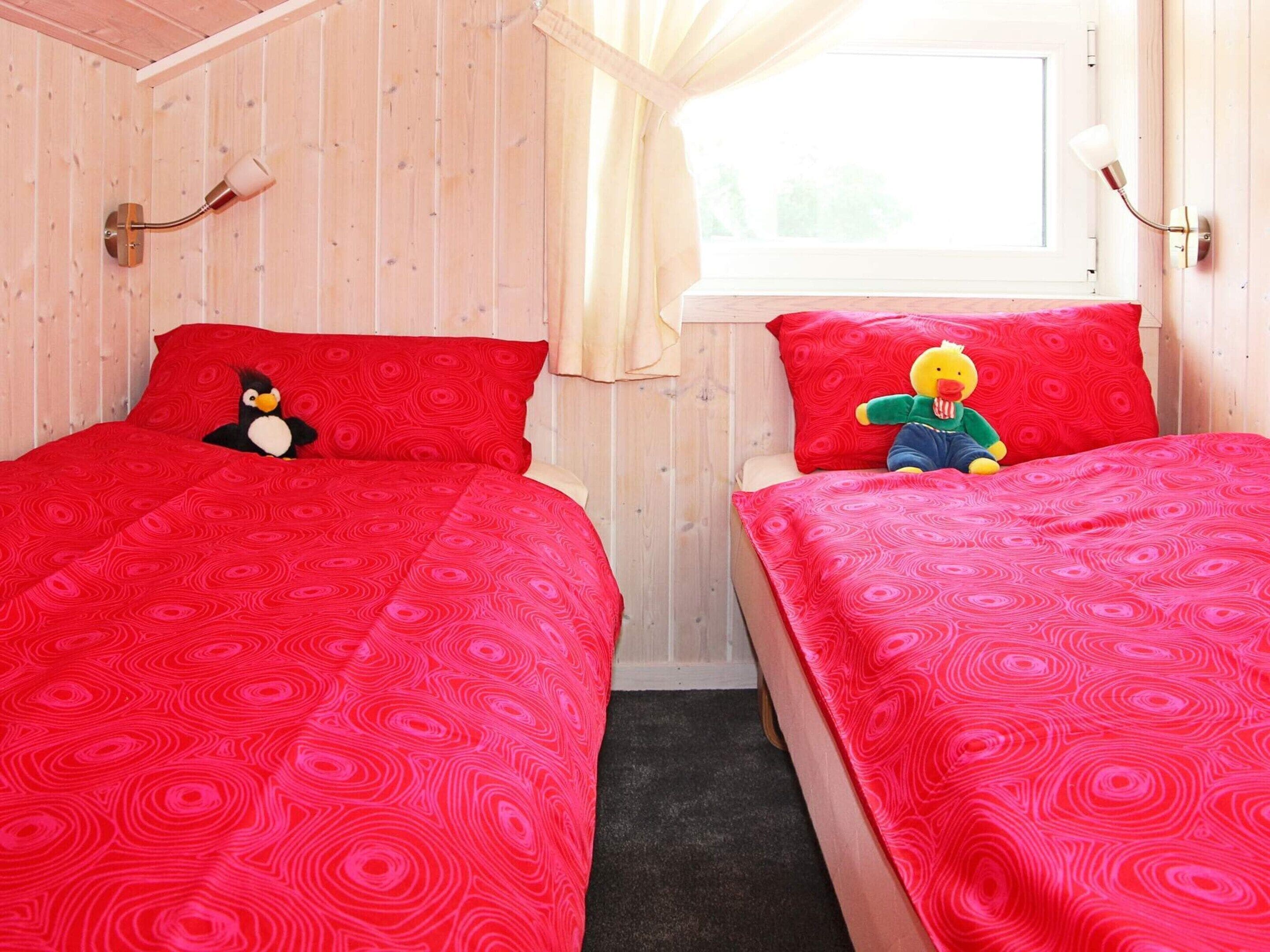 A room with two beds, each with a red patterned bedspread and two pillows, one with a penguin plush toy and the other with a duck plush toy.