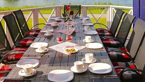 An outdoor dining table set with chairs, plates, cups, and a rose centerpiece, overlooking a lake with houses in the background. - Holiday Home in Otterndorf (Otterndorf)