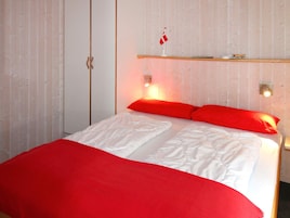 A bed with white bedding and red pillows, a wooden shelf above, and a wall with vertical lines.