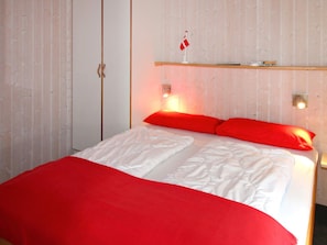 A bed with white bedding and red pillows, a wooden shelf above, and a wall with vertical lines. - Holiday Home in Otterndorf (Otterndorf)