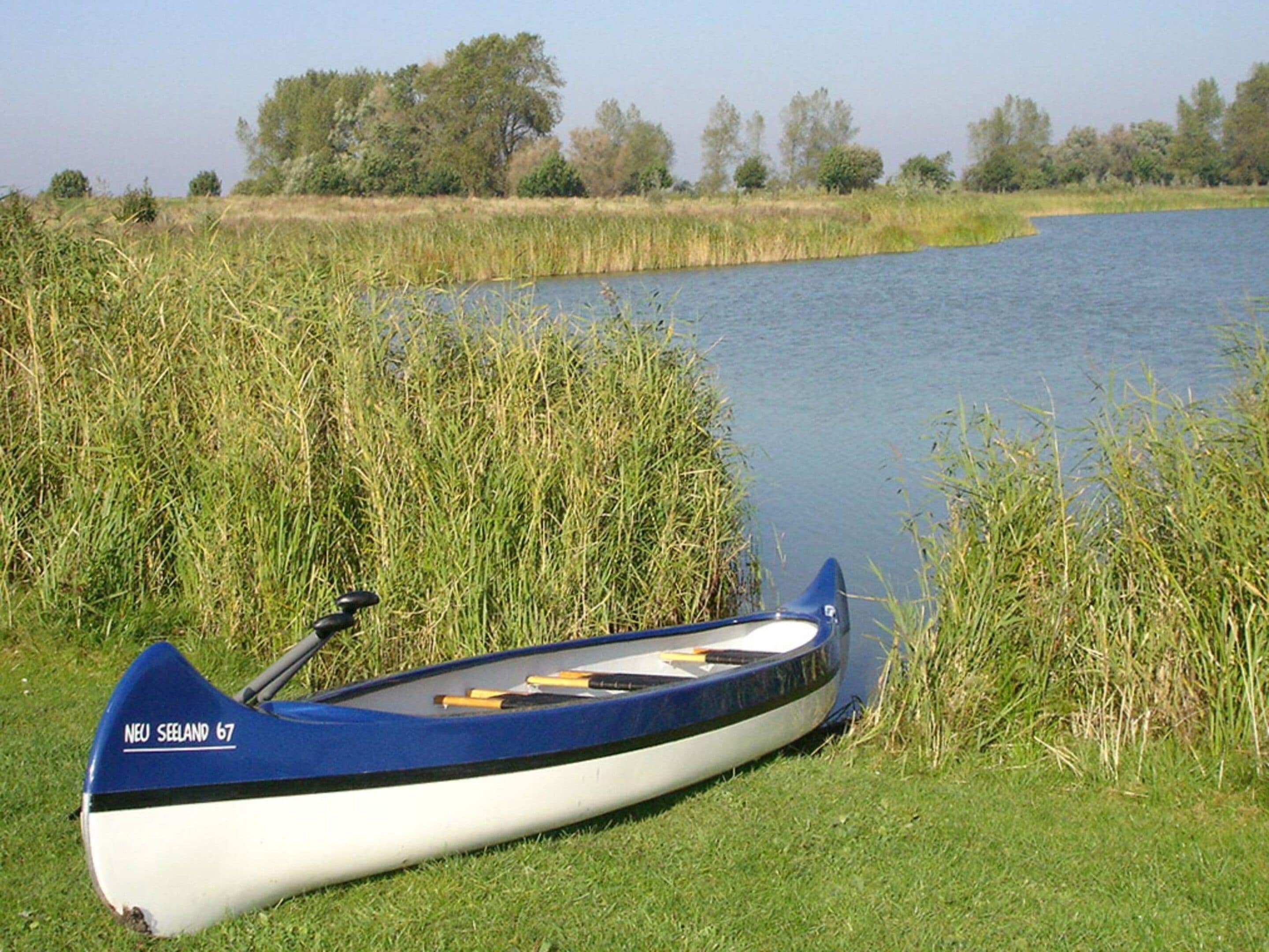 A canoe with the inscription 'NEW SEELAND 67' resting on grass beside a calm body of water.