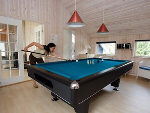 Mesa de billar en una habitación con paredes y techo de madera, una puerta que da al exterior y dos lámparas colgantes.