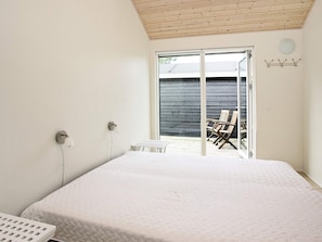 A bedroom with a bed, a small table, and a chair, a wall-mounted light fixture, and a sliding glass door leading to an outdoor seating area.