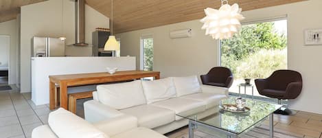 Un salon moderne avec une table basse en verre, un canapé blanc et des fauteuils foncés. On aperçoit une table à manger en bois à l’arrière-plan.