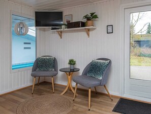 A room with two grey armchairs, a small round table, a television on a shelf, and a view of a swimming pool through a window. - 14 Person Holiday Home in Orsted (Ørsted)