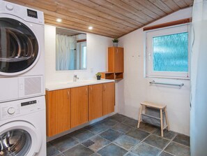 A compact bathroom with a washing machine, wooden cabinets, a small stool, and a window with a view of trees. - 14 Person Holiday Home in Orsted (Ørsted)