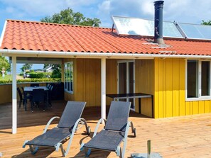 A covered outdoor area with a wooden deck, two lounge chairs, a table, and a yellow building with a red-tiled roof. - 4 Star Holiday Home in Nordborg (Nordborg)