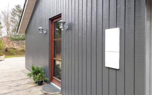 Un bâtiment à l’extérieur gris, avec une porte en bois et un petit porche agrémenté d’un pot de fleurs.