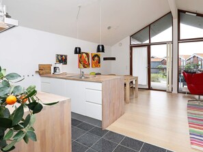 Una cocina moderna con una isla central, un área de comedor con una silla roja y vistas a una zona residencial a través de amplios ventanales.