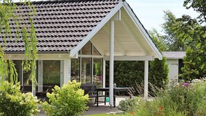 Un espace extérieur aménagé, abrité et doté de tables et de chaises, entouré d’une végétation luxuriante et de plantes en fleurs.