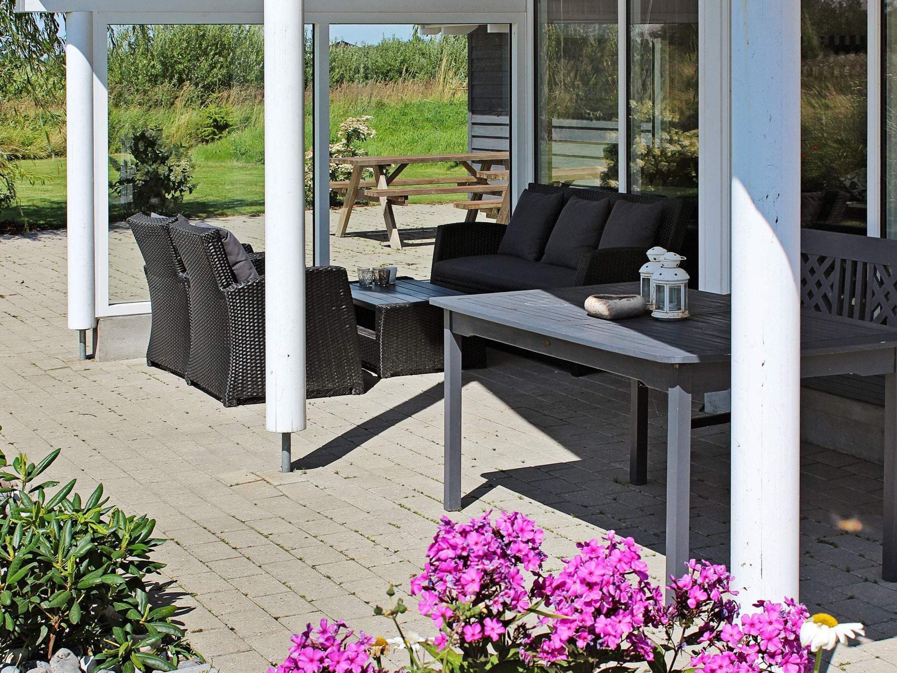 An outdoor seating area with wicker furniture and a table, surrounded by a wooden deck and a garden with flowers.