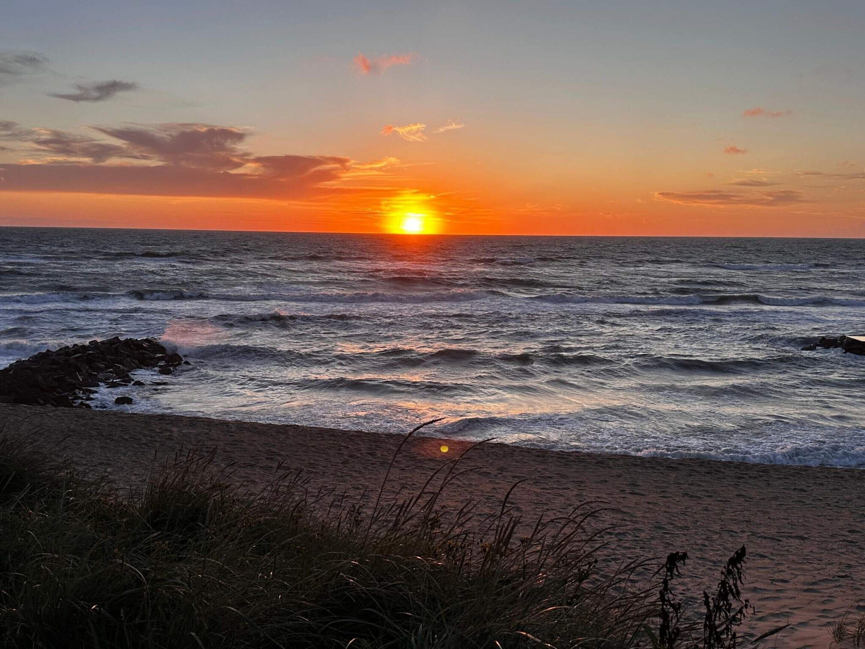 A sunset over the ocean with waves crashing onto a rocky shore.