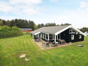 A black house with a covered patio, a lawn, and a red building in the background. - 4 Star Holiday Home in Hjorring (Hjørring)