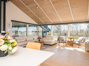 A spacious living area with a large sofa, a coffee table, and a dining table. There are large windows providing a view of the outdoors. - 4 Star Holiday Home in Bindslev (Bindslev)