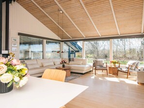 Amplia sala de estar con un sofá grande, una mesa de centro y una mesa de comedor. Grandes ventanas ofrecen vistas al exterior.
