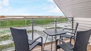Um pátio externo com mesa e cadeiras, com vista para um campo gramado e uma área industrial ao longe.