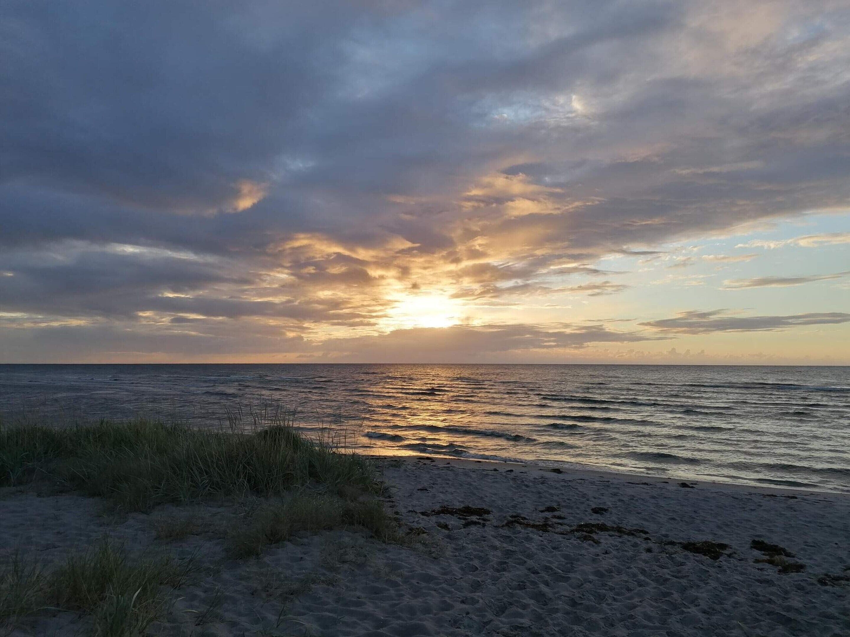 A beach with sand and grass, a cloudy sky, and the sun setting over the ocean.