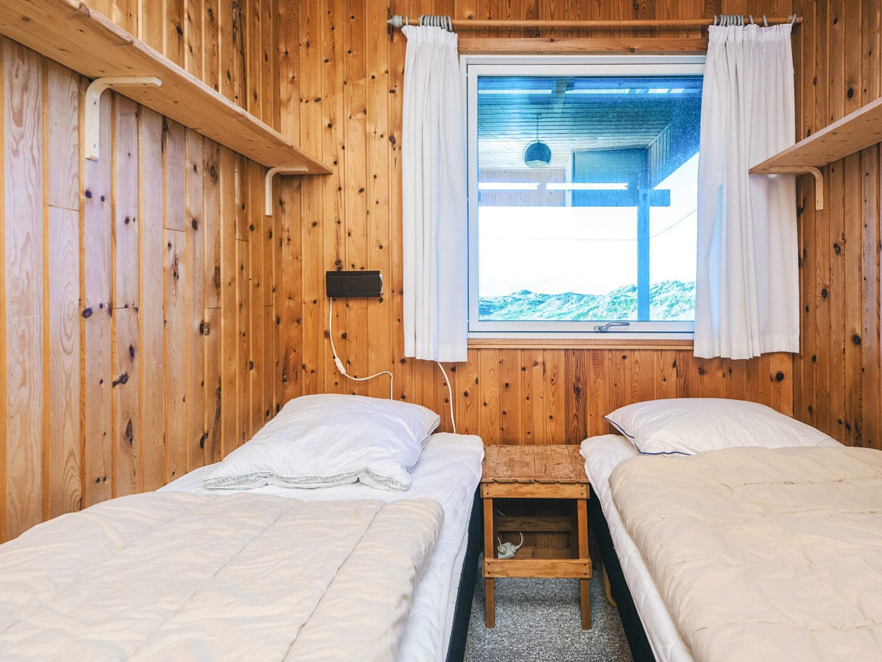 Ein Zimmer mit Holzboden und -möbeln, ausgestattet mit zwei Betten, einem Fenster mit Vorhängen und einem kleinen Holztisch.