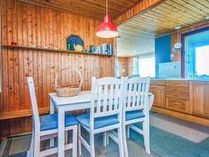 Interior de madera con una mesa de comedor, sillas, una lámpara colgante roja y una zona de cocina con un refrigerador.