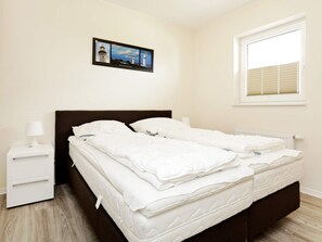 A hotel room with a bed, a nightstand, a lamp, a window with blinds, and a picture on the wall. - Holiday Home in Grossenbrode (Grossenbrode)