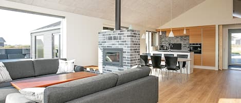 A modern living room with a fireplace, grey sofas, a wooden table, and a kitchen area with wooden cabinets and a black countertop.