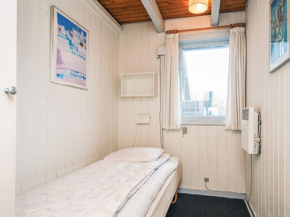 A small, single-bed room with a wooden ceiling, a window with curtains, a wall-mounted air conditioner, and a poster on the wall.