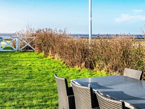 An outdoor seating area with a table and chairs, a white fence, and a view of the sea.