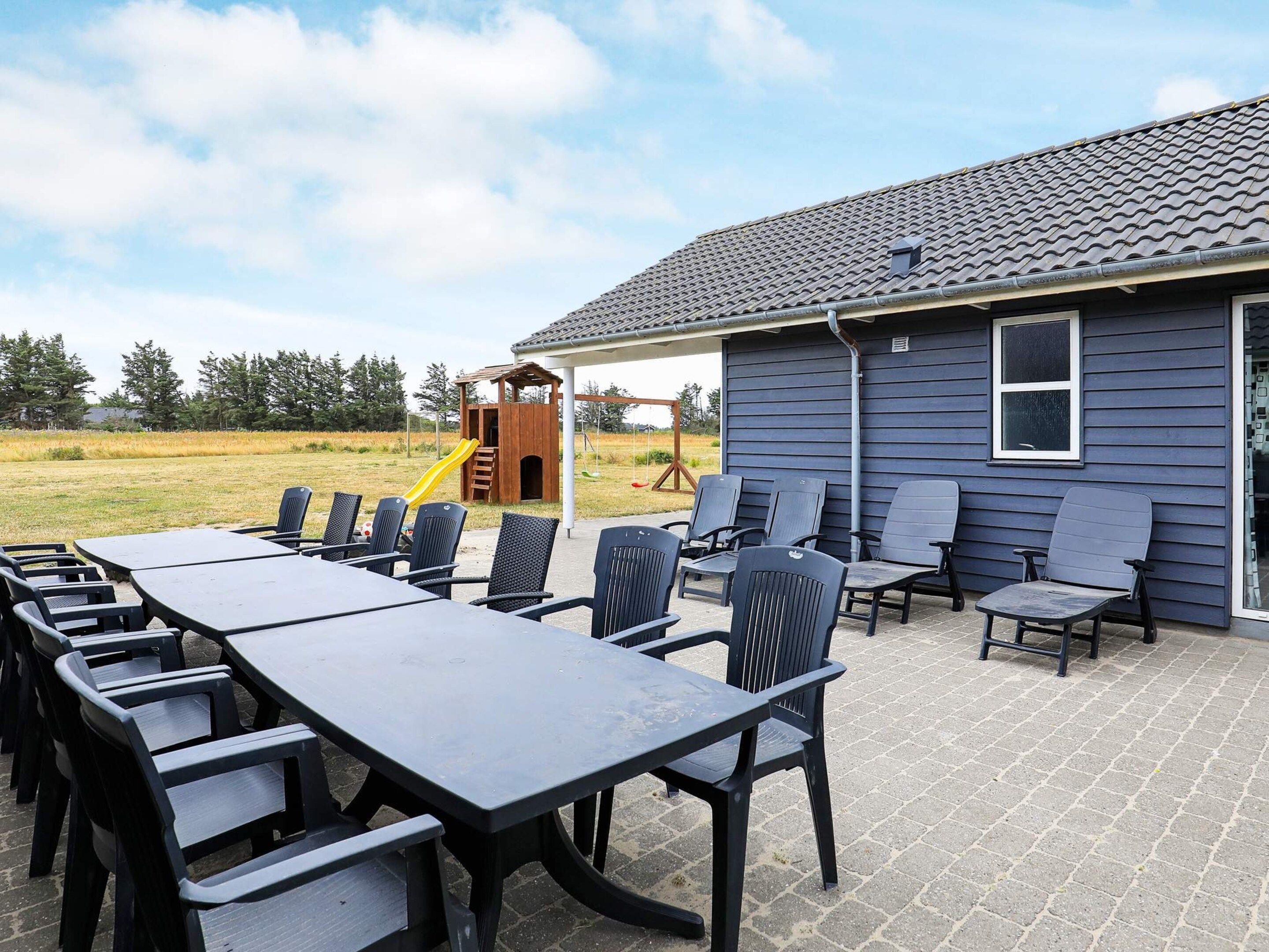 Une terrasse extérieure avec une longue table et des chaises, une structure de jeux et une maison dont le revêtement extérieur est de couleur sombre.