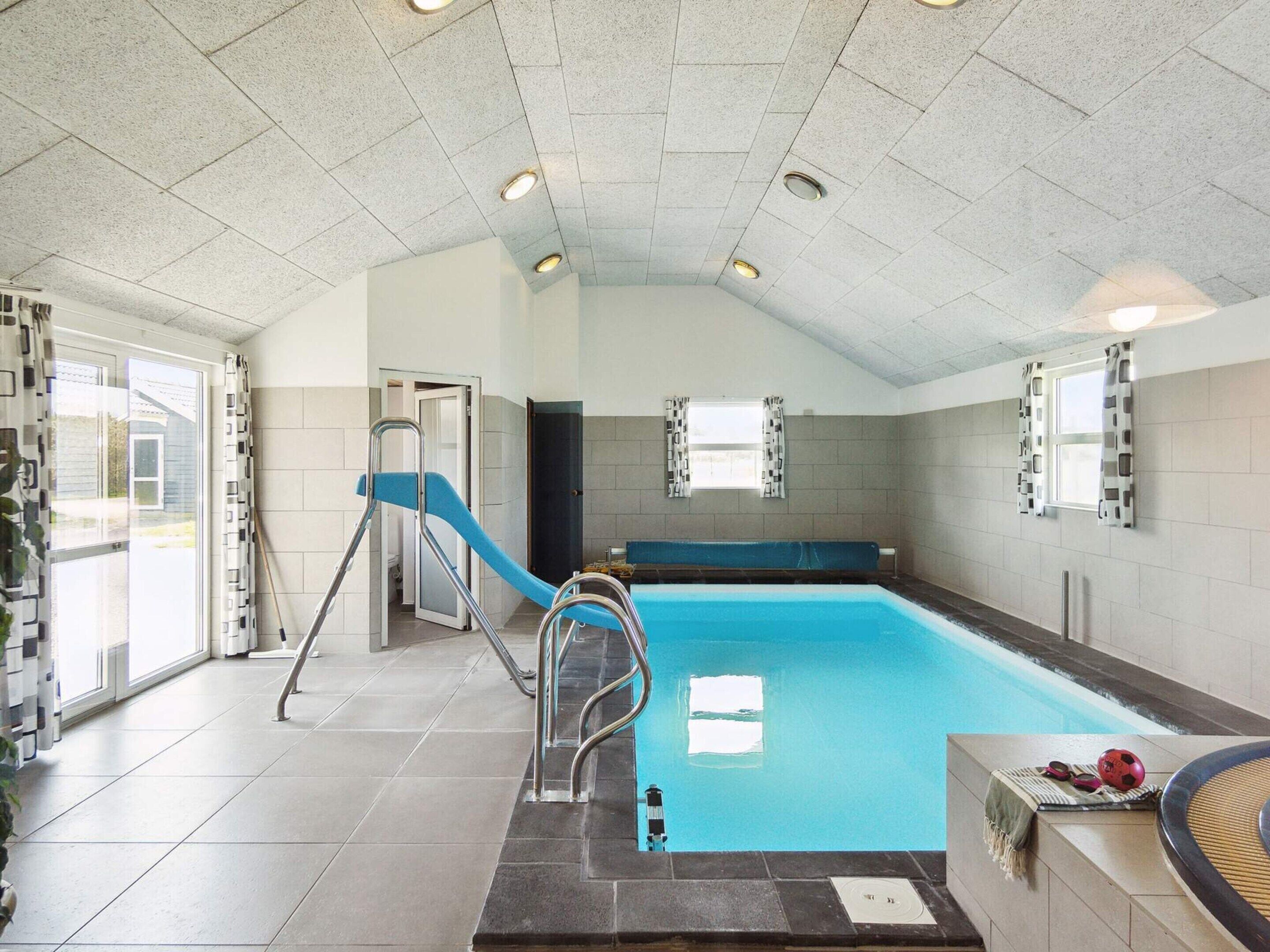 En indendørs swimmingpool med blå rutsjebane, en stige og et vindue med gardiner.