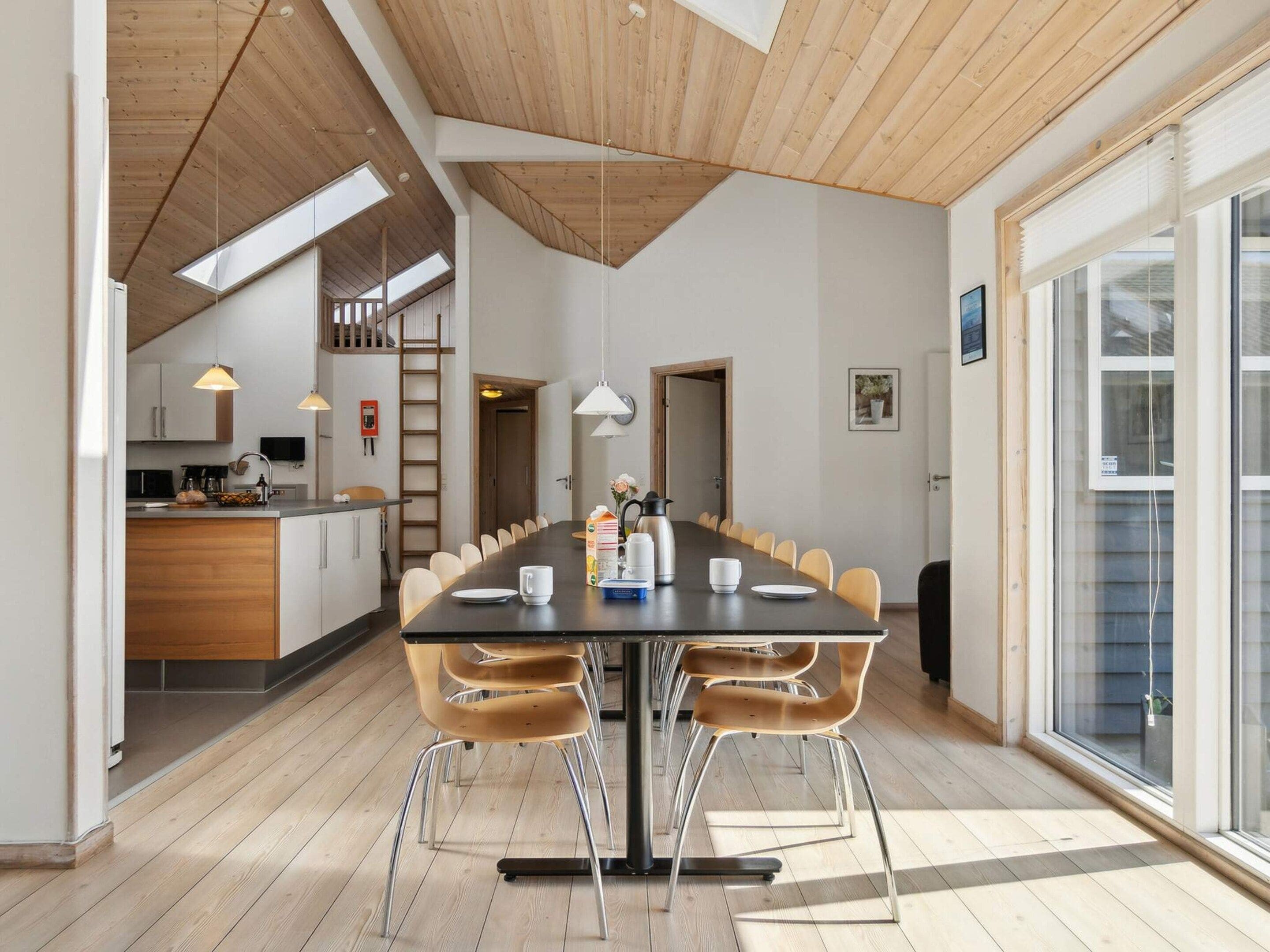 Et moderne køkken med træloft, et spiseområde med et langt bord og stole, samt et stort vindue der giver naturligt lys.