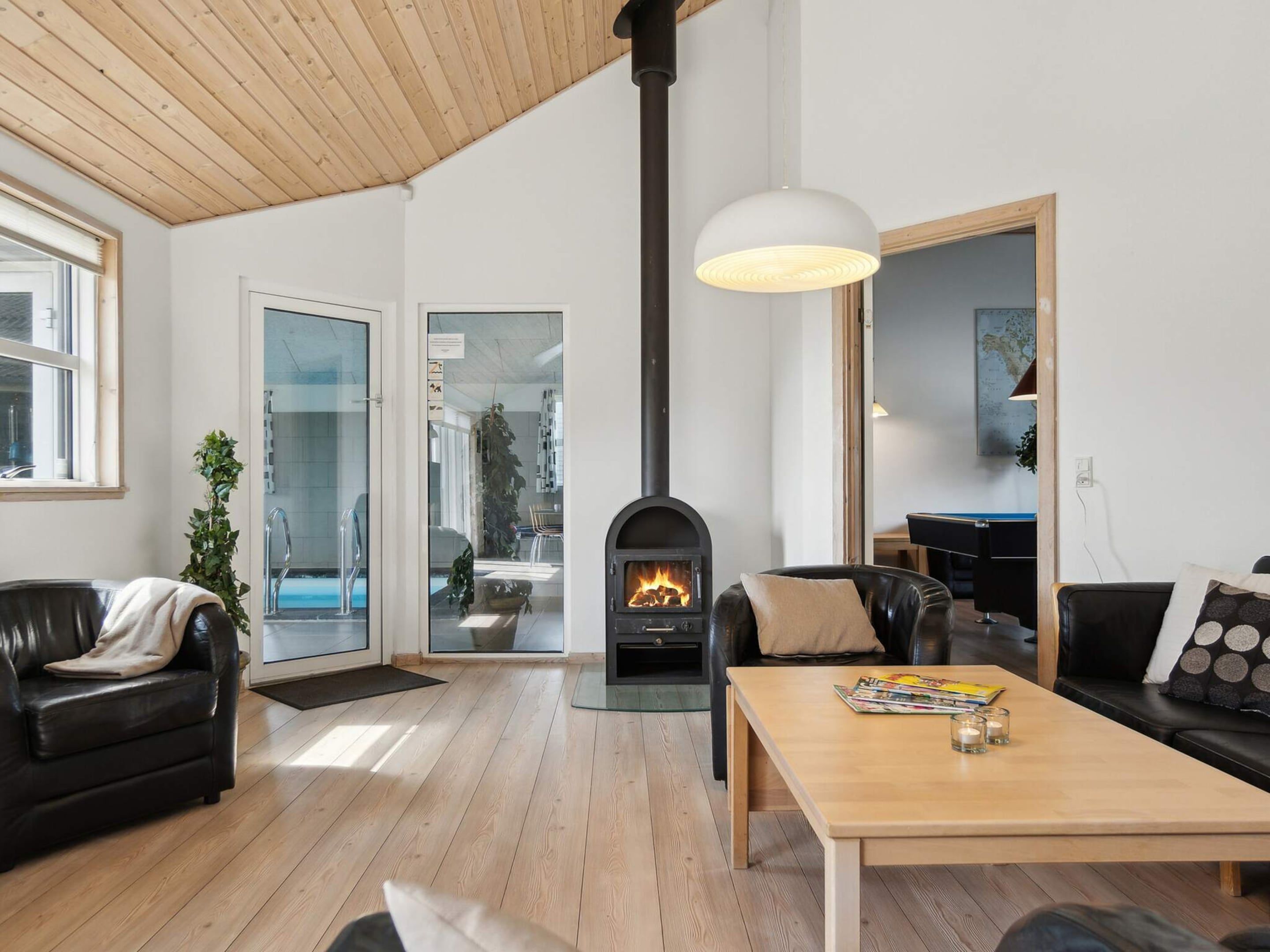 A modern living room with a fireplace, leather sofas, a wooden coffee table, and a view of an indoor pool.
