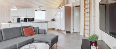 Un intérieur moderne avec un canapé gris, une table basse ronde et un coin cuisine doté de meubles blancs et d’un plafond en bois.