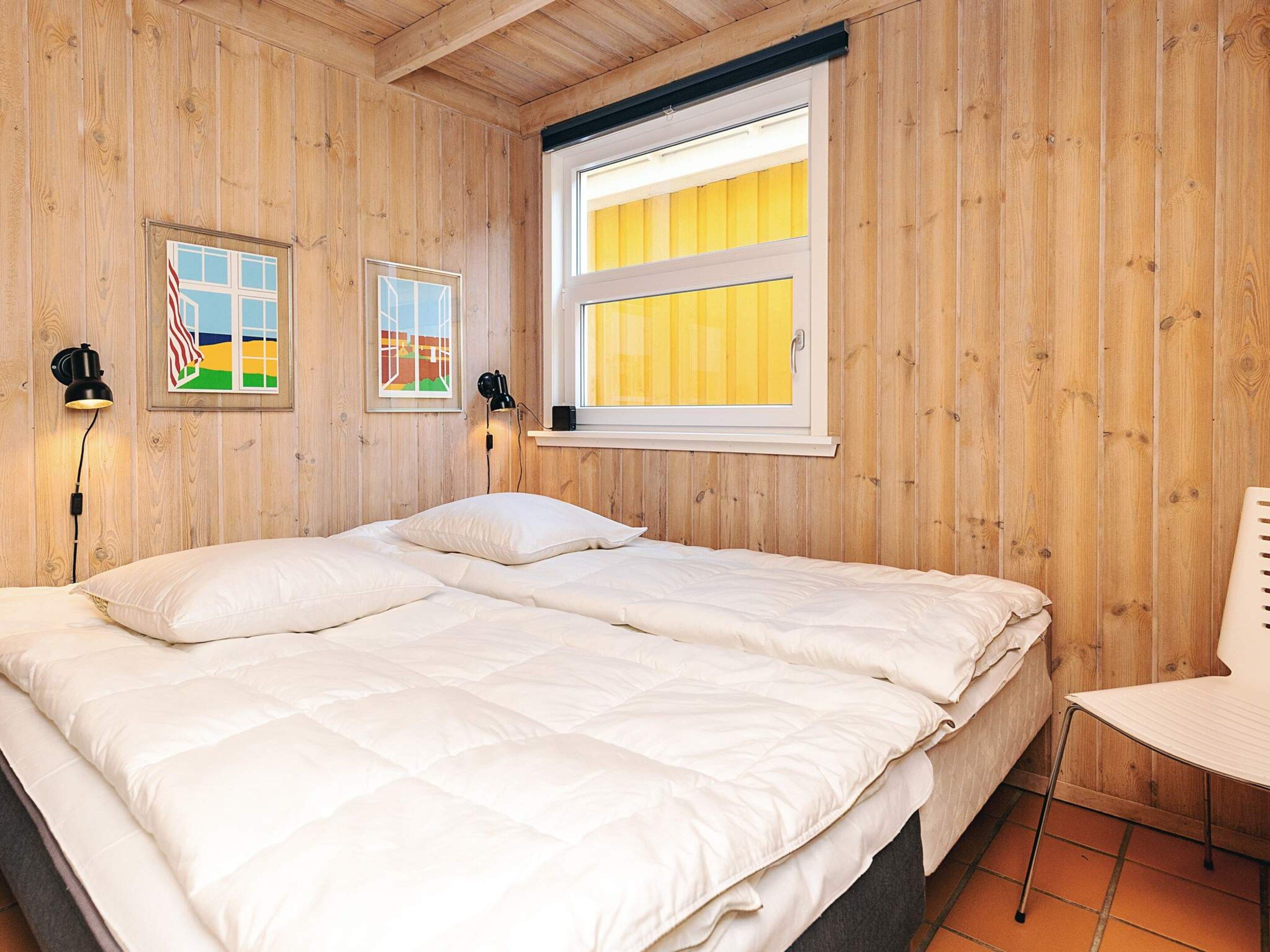 A wooden-paneled room with a large bed, two wall-mounted lamps, and a window with yellow blinds.