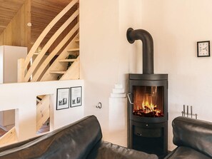 A modern living room with a black wood-burning stove, a wooden staircase, and a white wall with framed pictures.