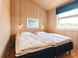 Une pièce aux murs en bois, un lit avec une literie blanche, deux lampes fixées au mur, un tableau encadré et une fenêtre donnant sur un paysage.