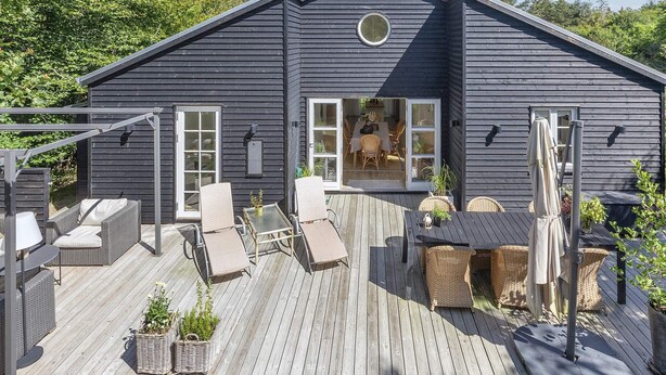 A dark wooden house with a deck, outdoor furniture, and a dining area visible through the open door. - 6 Person Holiday Home in Blokhus-by Traum (Blokhus)