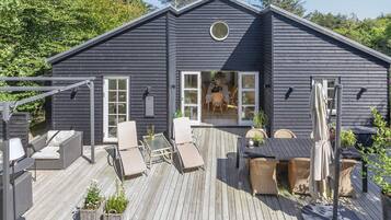 Una casa de madera oscura con un porche, muebles de exterior y un comedor visible a través de la puerta abierta.