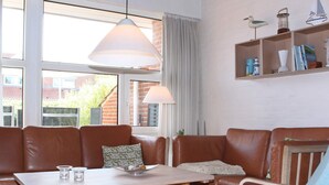 Un salon chaleureux avec un canapé en cuir marron, une table basse en bois et un fauteuil bleu. Une fenêtre offre une vue sur un bâtiment en briques à l’extérieur.