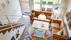 Un salon spacieux avec un canapé en cuir marron, une table basse et un tapis coloré. Un escalier se trouve sur la gauche, et de grandes fenêtres offrent une vue sur un jardin.