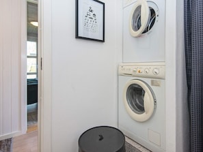 A laundry room with two front-loading washing machines, a framed picture on the wall, and a small trash bin.